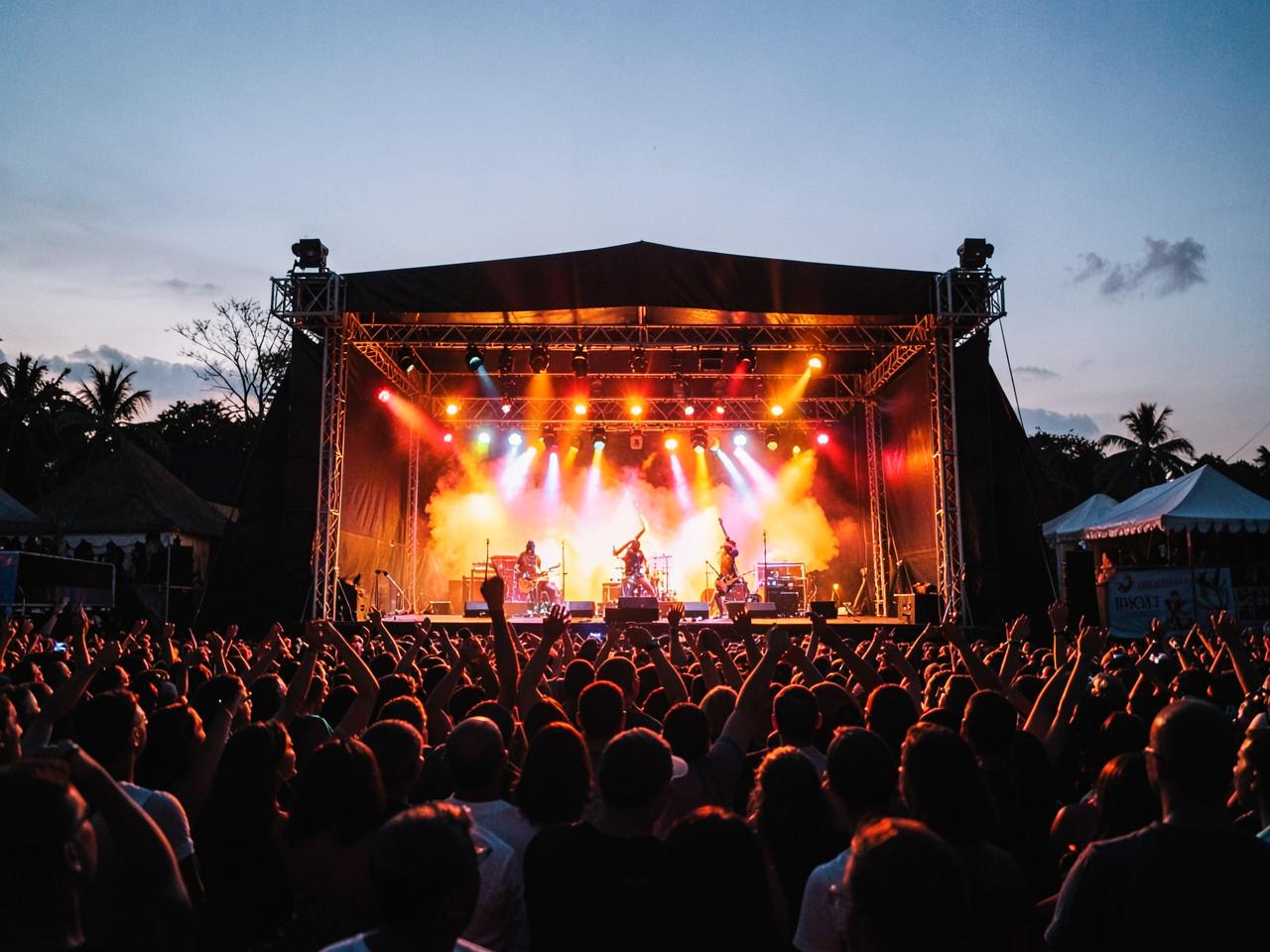 Outdoor festival stage at dusk with warm lights and a crowd with raised hands